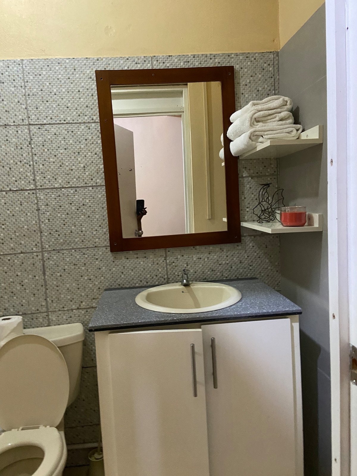 A compact bathroom features gray tiled walls and a mirror above a white sink. A small white cabinet with two doors provides storage. Towels are neatly arranged on a shelf, and a lit candle adds a touch of detail to the space.