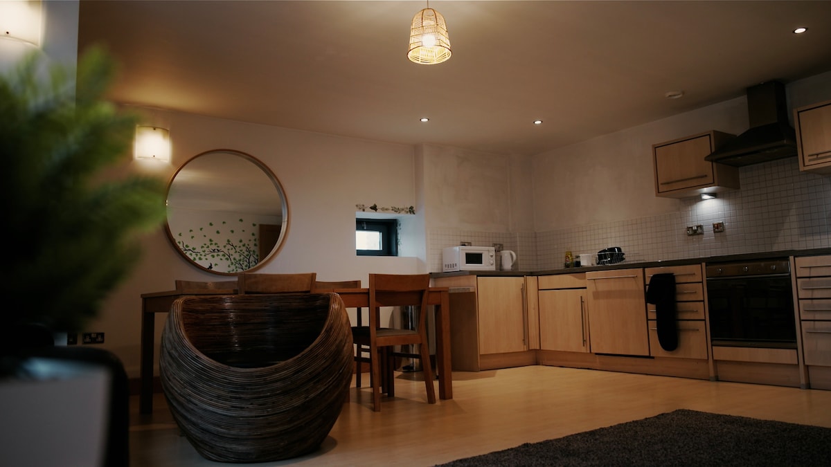 The kitchen and dining area features wooden cabinetry and modern appliances. A round mirror reflects light, enhancing the space. A decorative bowl and a dining table with four chairs are visible, while soft lighting creates a comfortable environment. A plush rug is positioned on the floor.