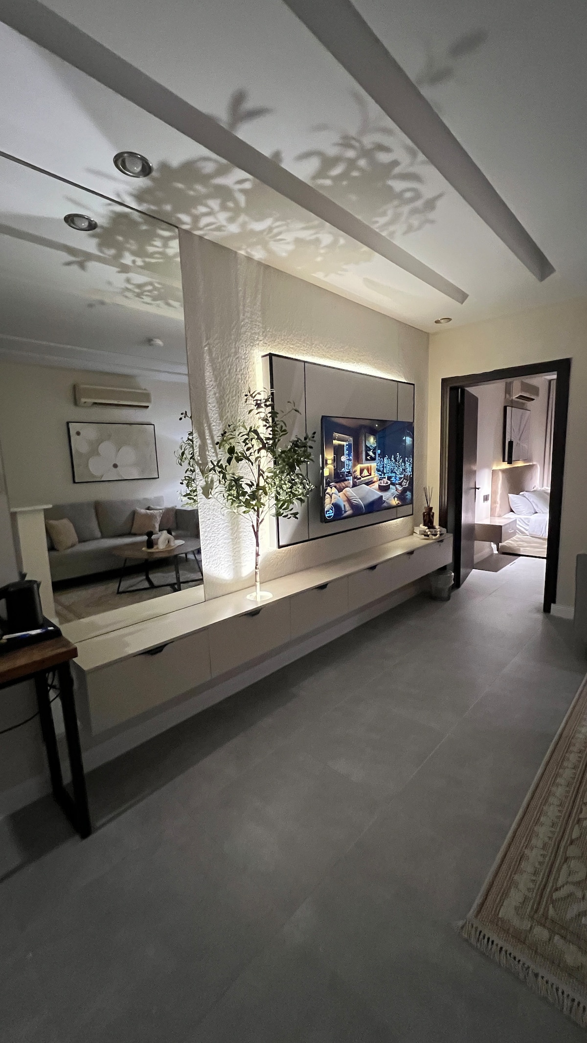 A modern living area is visible, featuring a wall-mounted television framed by softly glowing lighting. A decorative plant adds a touch of greenery to the room. A cozy seating area is positioned in the background, with a doorway leading to a bedroom area.