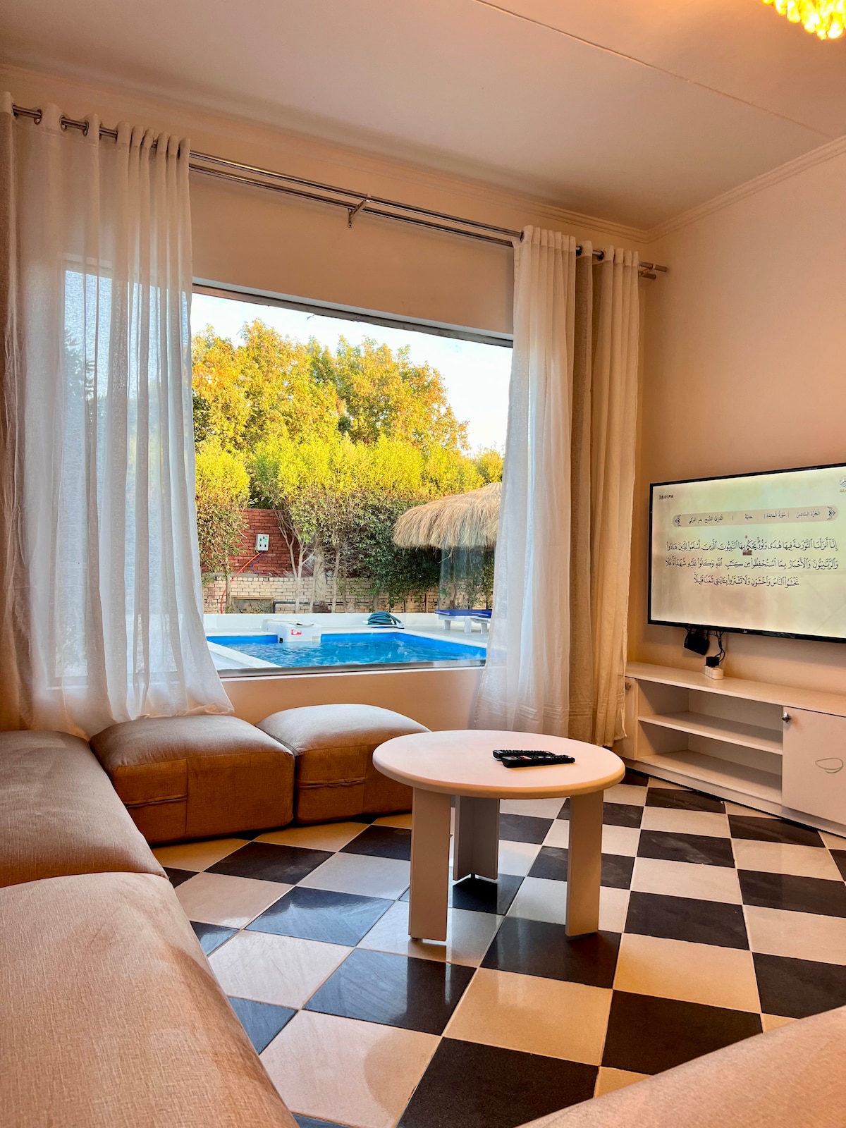 A cozy living area features a large sectional sofa and a circular coffee table. Natural light filters through floor-to-ceiling curtains, illuminating the checkered floor. A clear view of the outdoor area reveals a swimming pool surrounded by greenery.
