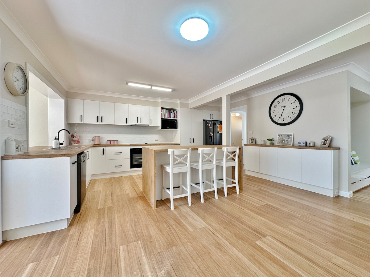 The spacious kitchen features sleek white cabinetry and a central island with seating for four. Modern appliances, including a refrigerator and oven, are visible. Natural light fills the area, highlighting the warm wooden floor, while a large clock adorns the wall for easy timekeeping.