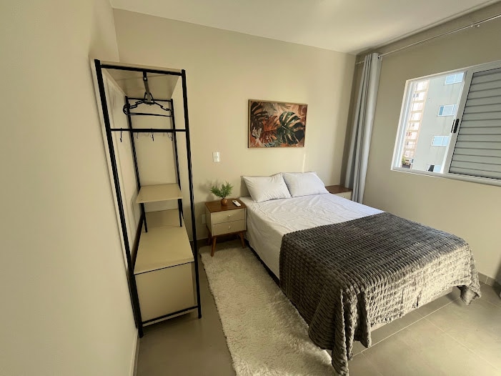 A bright bedroom features a spacious bed with a neutral-toned blanket. A light-colored rug adds warmth to the tiled floor. A minimalist wardrobe stands next to the bed, equipped with hanging space. Natural light enters through a nearby window with simple blinds.