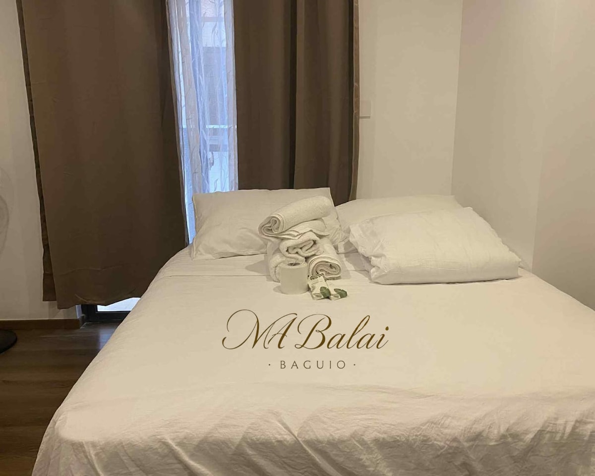 A spacious queen bed is positioned against a simple wall, topped with white linens and neatly rolled towels. Soft brown curtains frame a window, allowing natural light to fill the serene space, while a light wooden floor complements the neutral color palette.