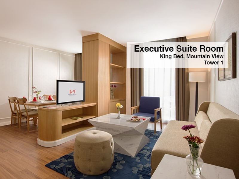The Executive Suite Room features a seating area with a spacious sofa and armchair, complemented by a marble coffee table. A wooden desk with chairs is positioned near a large window, allowing natural light to illuminate the space. A television is mounted on the wooden cabinetry.