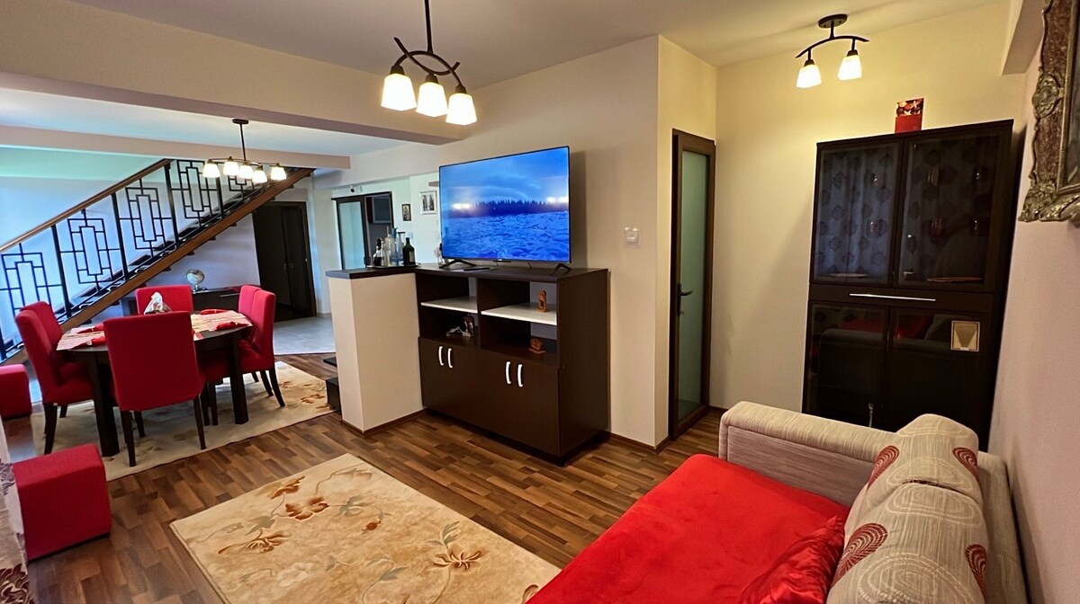 A spacious living area is presented, featuring a large television mounted on a dark cabinet. A beige sofa with a red accent pillow sits beside a patterned area rug. A dining area is visible in the background, equipped with a round table and red chairs, under soft lighting.