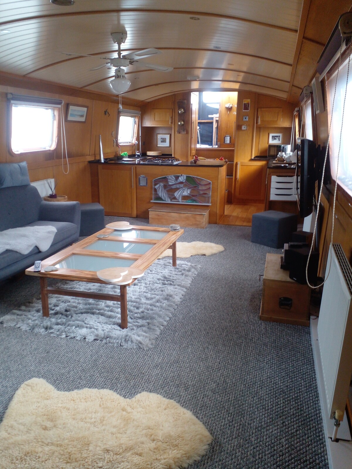 The interior of the boat features warm wooden paneling and a spacious layout. A comfortable sofa is positioned near a coffee table, and a well-equipped kitchen can be seen in the background. Textured rugs add softness to the flooring, while large windows provide ample natural light.