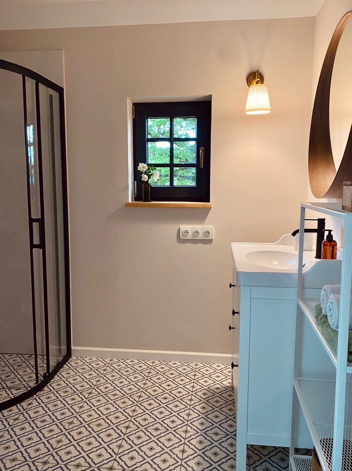 A well-appointed bathroom features a walk-in shower enclosed by glass. Natural light enters through a small window adorned with a potted plant. A white vanity hosts a sink and is complemented by a round mirror, enhancing the minimalist design.