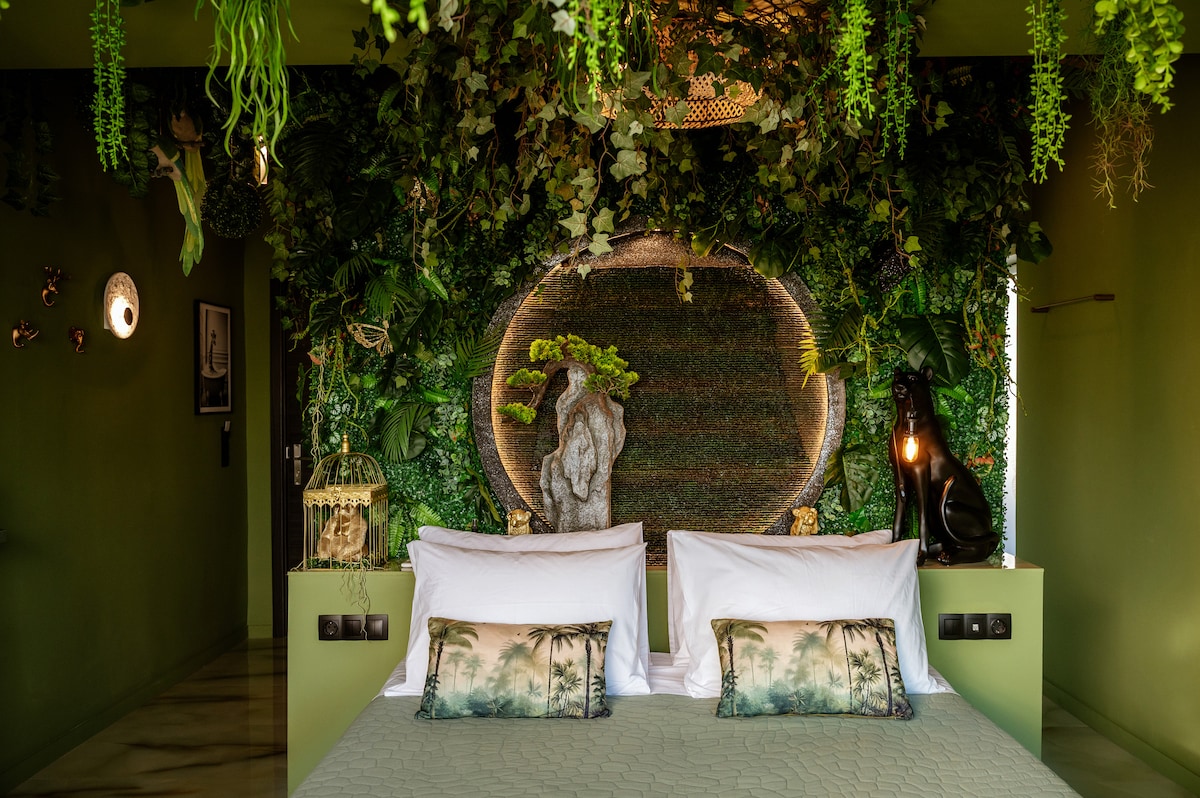 A spacious bed is layered with textured pillows against a backdrop of lush greenery and natural elements. Decorative lighting and artful pieces, including a stone figure, enhance the organic aesthetic of the room, creating a serene and inviting atmosphere.