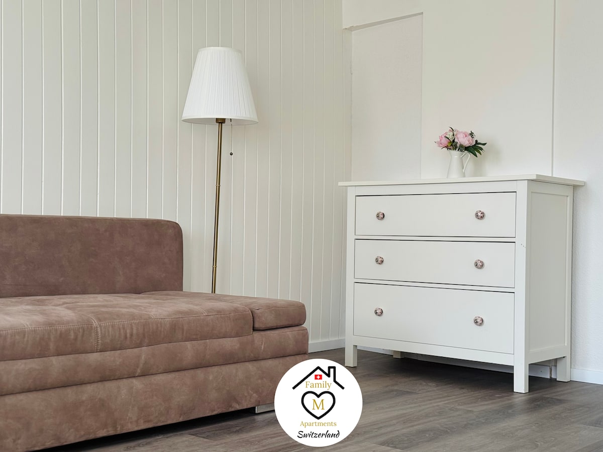 A spacious living area features a modern light fixture beside a cozy, brown sectional sofa. A sleek, white dresser with three drawers is positioned against the wall, adorned with a small vase of pink flowers, bringing a touch of color to the room.