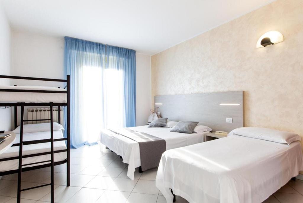 A light-filled room features two spacious beds and a bunk bed against the wall. Soft blue curtains frame the large window, while minimalistic light fixtures above the headboard provide gentle illumination. Neutral tones create a calming environment, inviting relaxation.