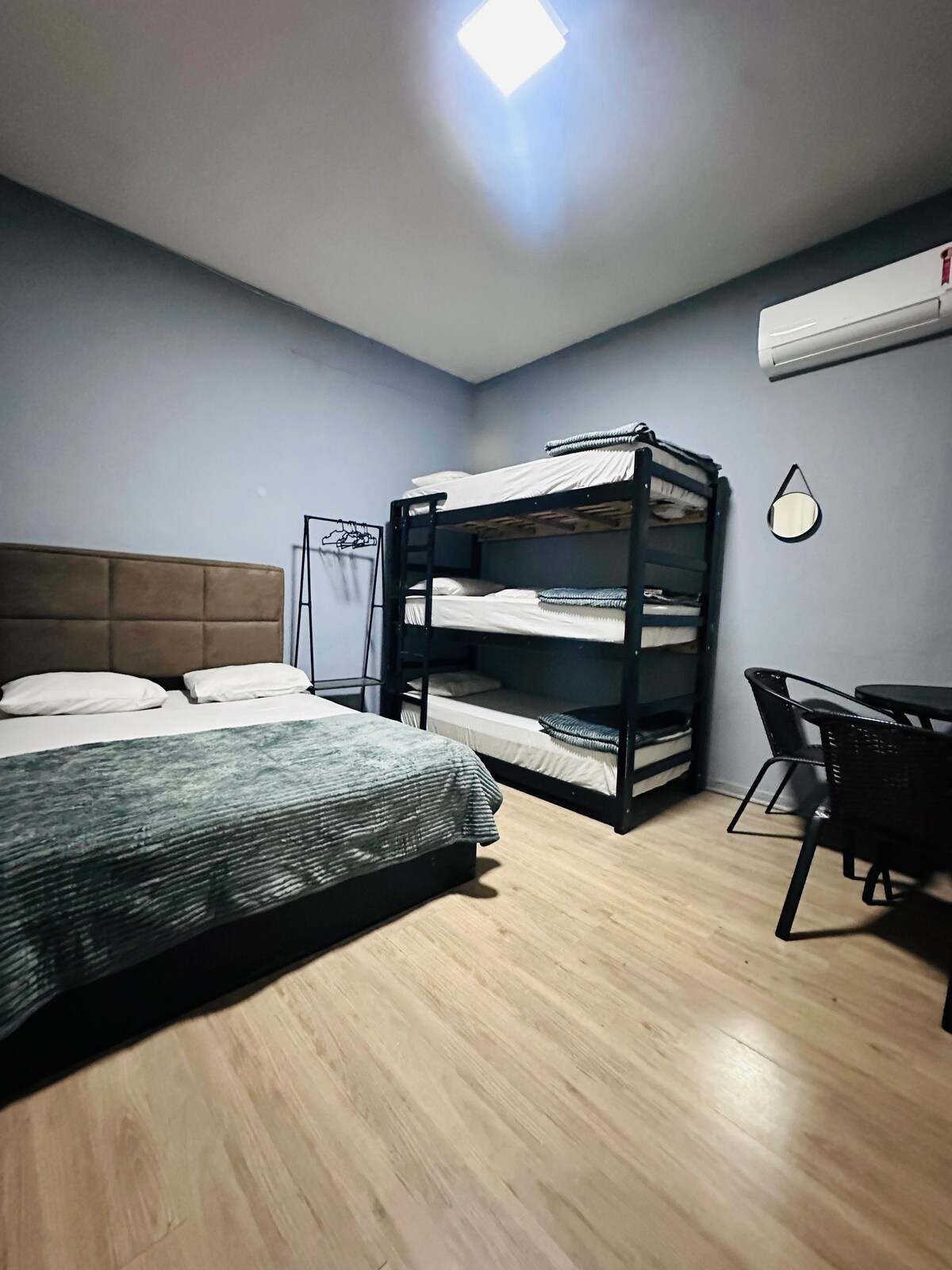 A studio room features a spacious double bed alongside a triple bunk bed. A small dining table and chairs are positioned near the window. Air conditioning is mounted on the wall, and a coat rack is visible in the corner, with soft lighting illuminating the space.