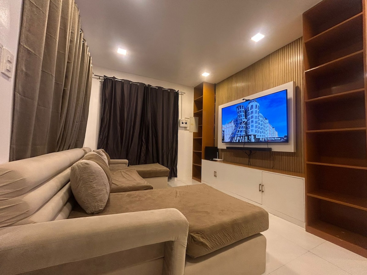 A spacious living room is furnished with a neutral-colored sectional sofa and a flat-screen smart TV mounted on a wooden accent wall. Dark curtains frame the windows, providing a cozy atmosphere, while open shelving units offer storage space.