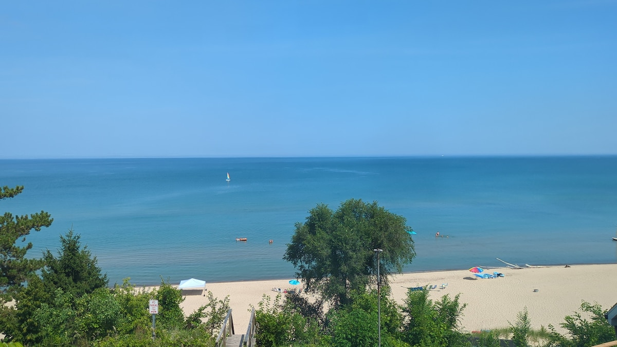A serene beach scene captures the calm blue waters extending to the horizon. Soft sandy shores are lined with green vegetation, and a few beach umbrellas dot the sandy area. A small sailboat can be seen gently gliding on the water.