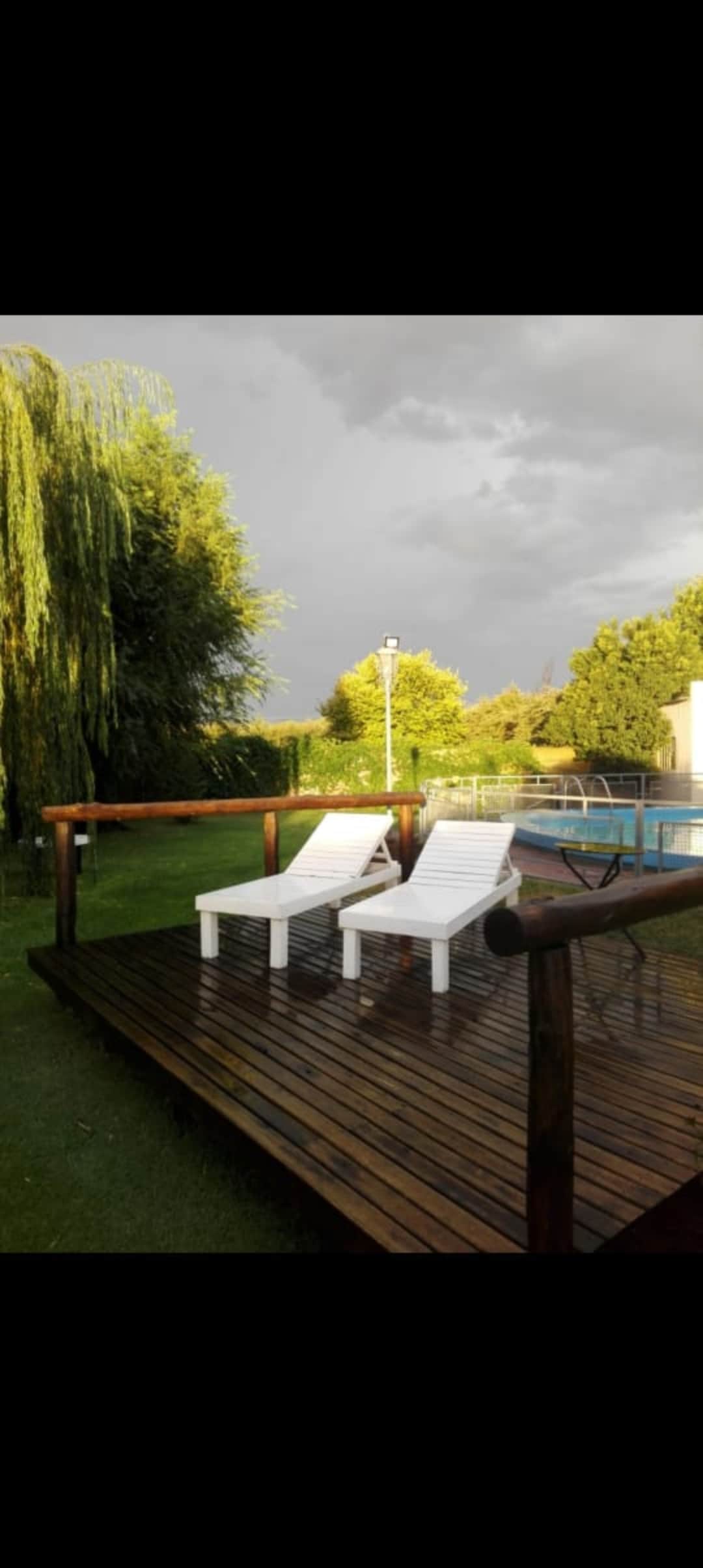 A wooden deck is shown with two white lounge chairs positioned for relaxation. Lush green trees surround the area, and an outdoor swimming pool is visible in the background. The sky appears overcast, creating a serene atmosphere.