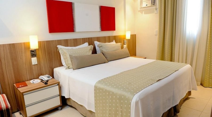 The room features a spacious bed with neutral bedding and decorative cushions, positioned against a wooden headboard. A bedside table with a phone is visible on one side, while soft lighting creates a calm environment. Light curtains frame a window allowing natural light into the space.
