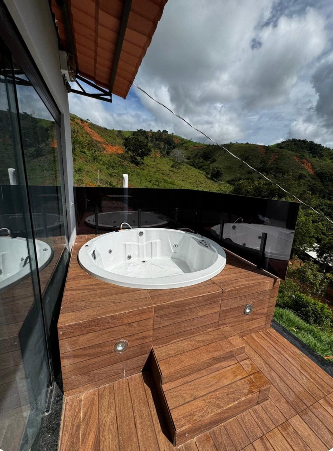An outdoor jacuzzi is positioned on a wooden deck, surrounded by lush green hills. The jacuzzi features several jets and is complemented by a stylish glass barrier that provides unobstructed views of the natural landscape. Stairs lead up to the jacuzzi area.