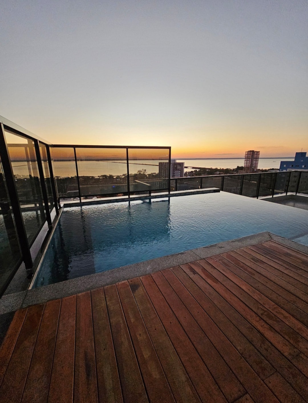 An infinity pool rests on a wooden deck, offering expansive views of the horizon during sunset. The glass railing frames the pool area, ensuring an unobstructed sight of the skyline and the water beyond, with colors ranging from warm orange to deep blue.