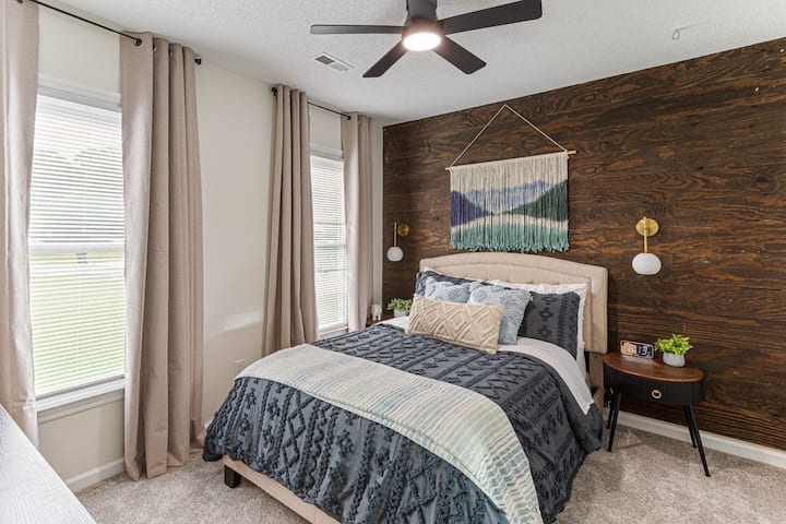 Warm and inviting bedroom with a full bed, rustic wood accent wall, and cozy layered bedding. Natural light fills the space, complemented by modern lighting and a peaceful ambiance.