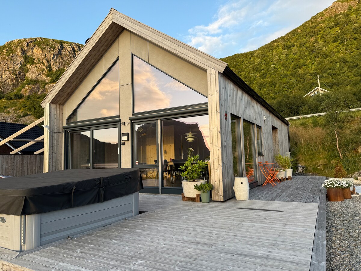 A modern cabin features large glass doors that provide an open view to the surrounding landscape. A deck area is adorned with potted plants and casual seating. The hot tub is positioned prominently, inviting relaxation amid natural surroundings.