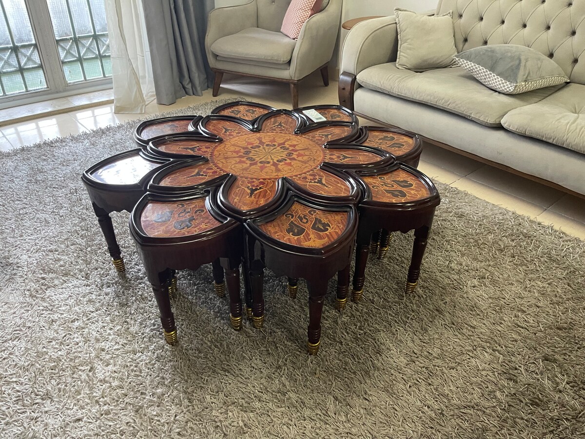 A unique coffee table with an intricate floral design is centered on a soft, beige rug. Surrounding the table, two light-colored armchairs provide comfortable seating. Natural light filters through the large window, accentuating the room's welcoming and open feel.