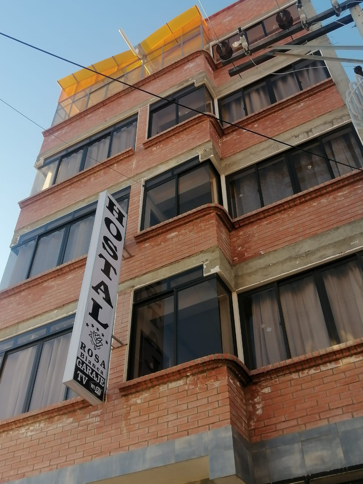 An exterior view of a four-story brick building is presented, featuring large windows with curtains that soften the natural light. A sign indicating 'HOSTAL' hangs prominently from the facade, while a yellow canopied rooftop offers a glimpse of outdoor seating. Power lines can also be seen overhead.