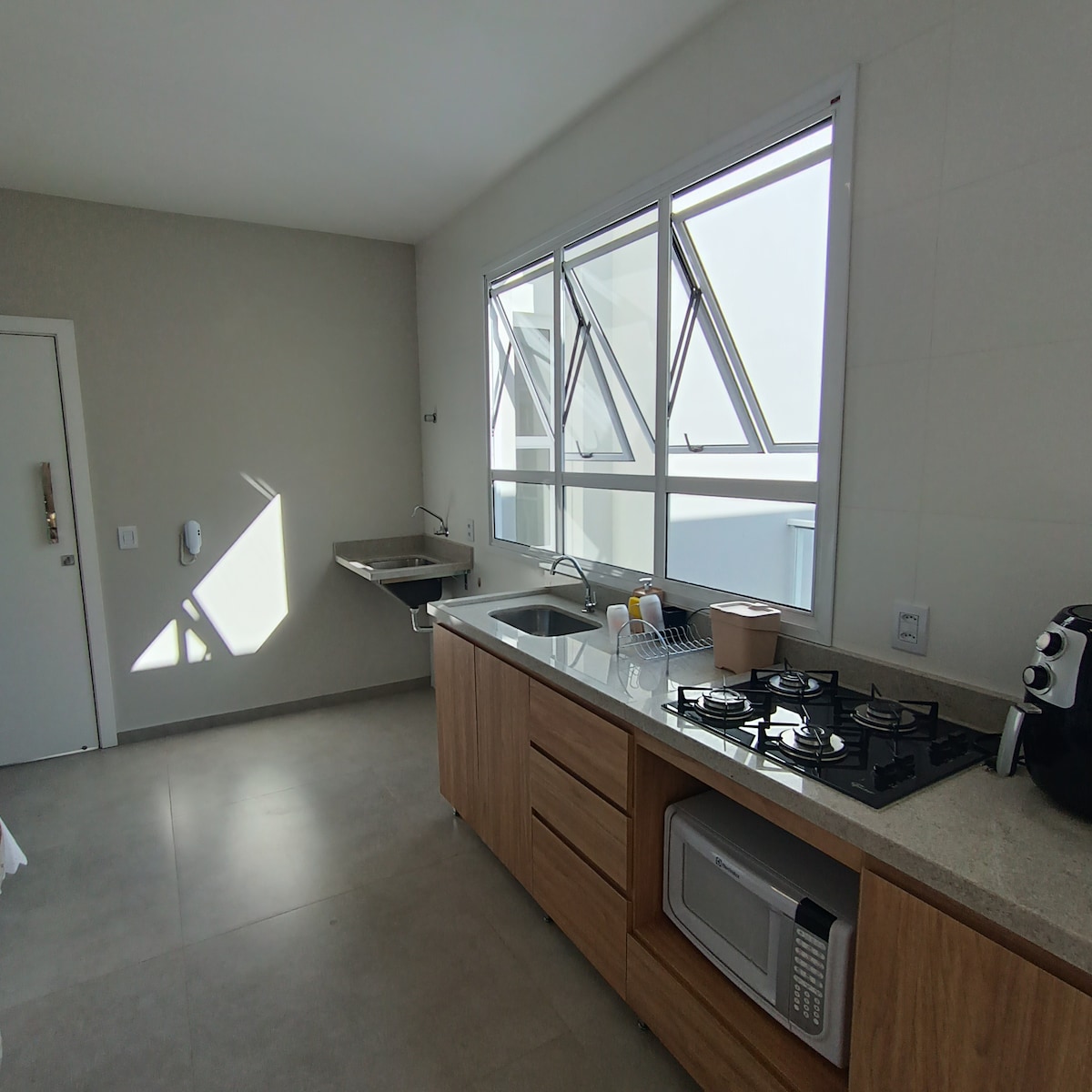 A bright kitchen area features large windows allowing natural light, with a modern layout. Wooden cabinetry holds a gas stove, a microwave, and a sink. Counter space accommodates kitchen essentials, while sleek surfaces maintain a clean aesthetic.
