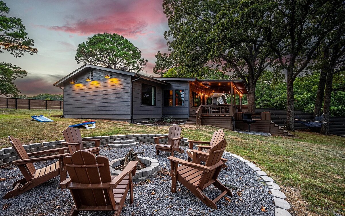 A cozy cabin is set against a colorful sky, surrounded by mature trees. A fire pit with wooden chairs is positioned in the foreground, providing a communal space. The cabin features a welcoming porch with soft lighting, enhancing the outdoor setting.