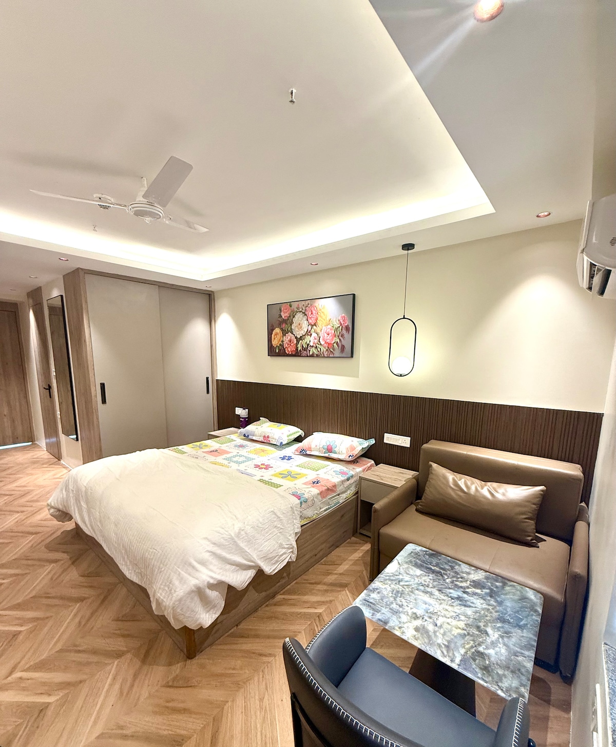 A contemporary room features a spacious bed adorned with a colorful quilt. A small seating area is present, complemented by a marble-topped table. Wall art depicting flowers adds a touch of color, while modern lighting brightens the stylish interior.