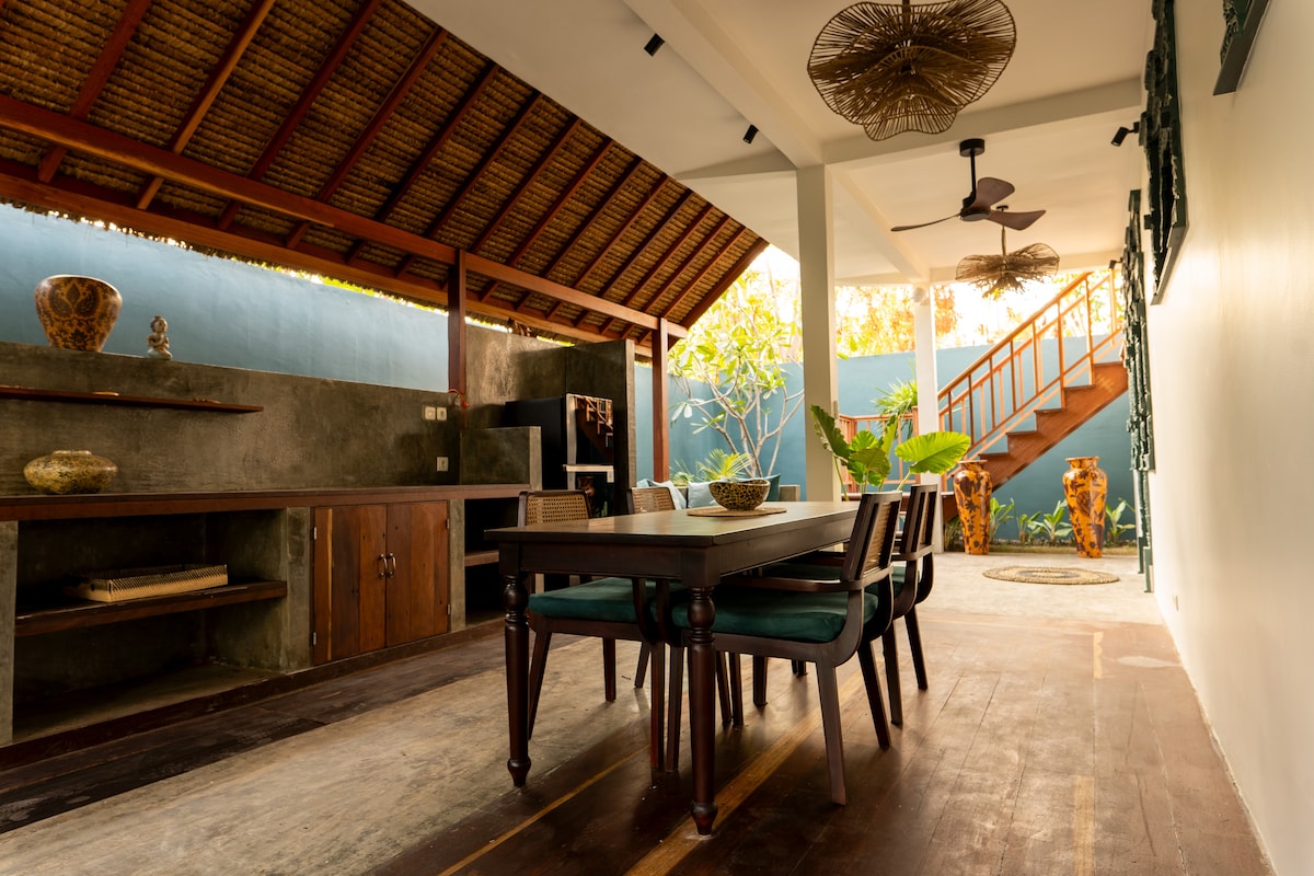 A dining area is showcased with a wooden table surrounded by dark green chairs. Natural light filters through an open space, highlighting a textured ceiling made of bamboo. An adjacent stairway is visible, leading to another level, with greenery placed thoughtfully in the background.