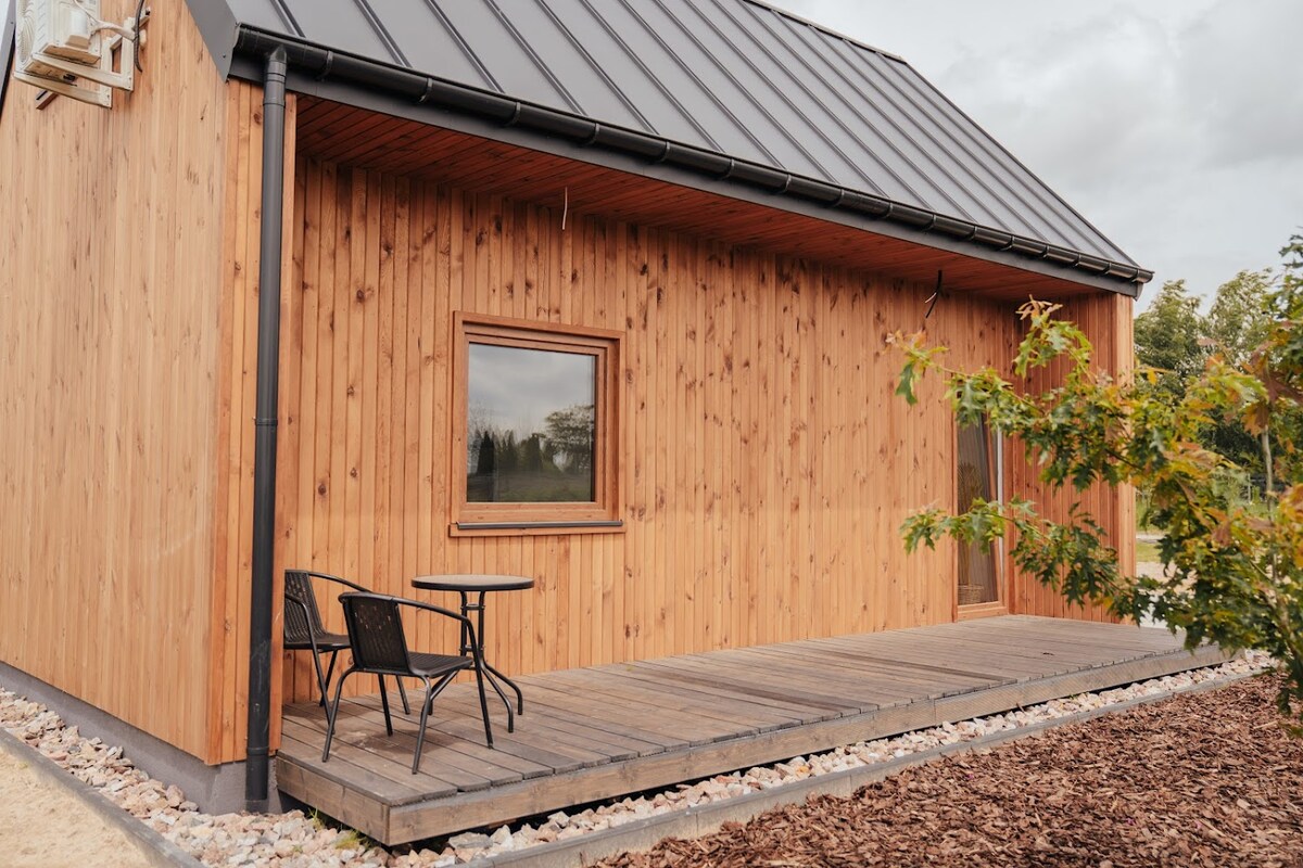 A wooden cabin features a small outdoor deck with two black chairs positioned near a square table. The natural wood siding complements the landscape, with a large window allowing for ample natural light. Stone landscaping surrounds the cabin, enhancing the serene outdoor setting.