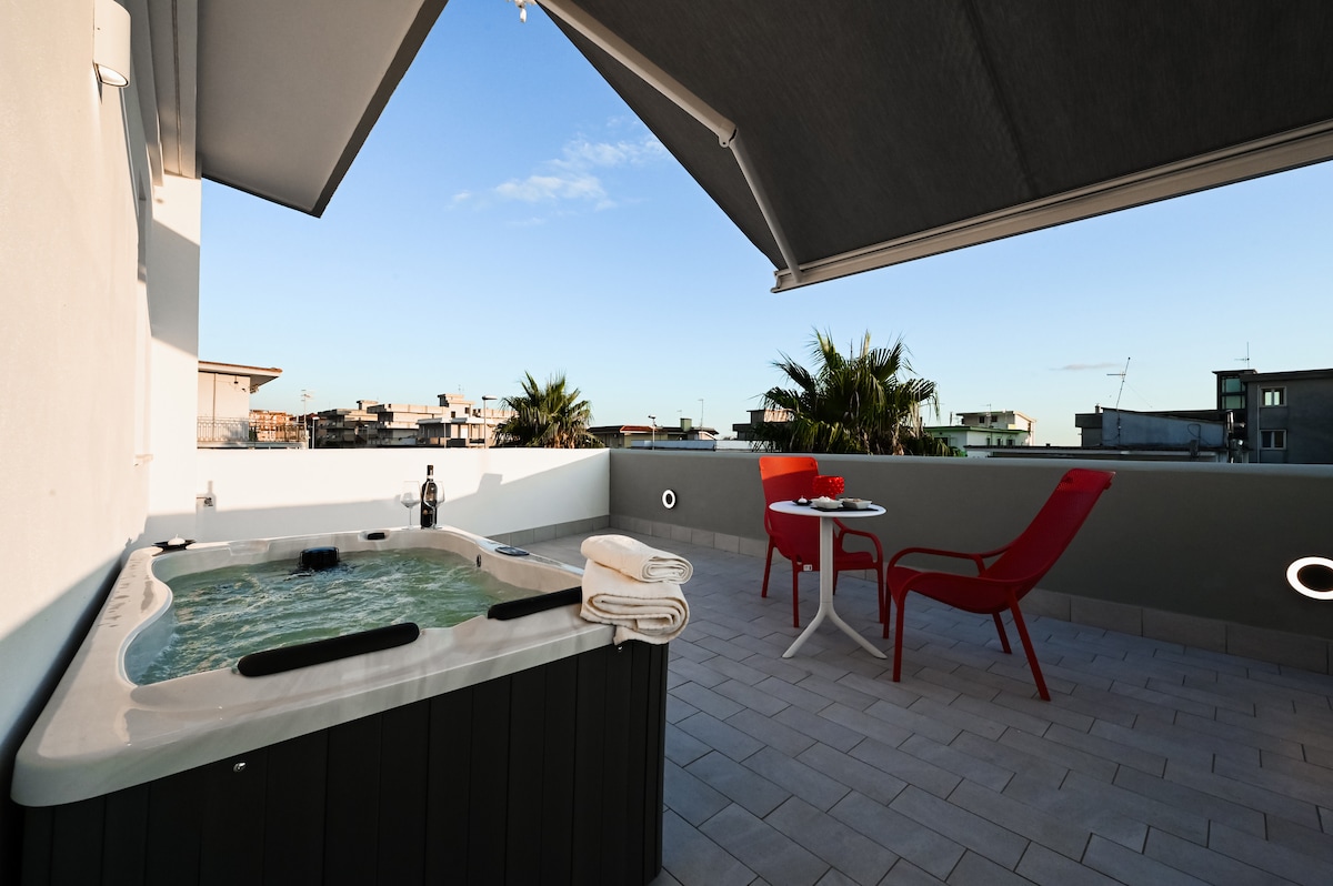 A private terrace features a jacuzzi surrounded by a low wall, with views of the skyline. Red chairs and a small table are positioned beside the jacuzzi, inviting relaxation. The space is brightened by natural light from the clear blue sky above.