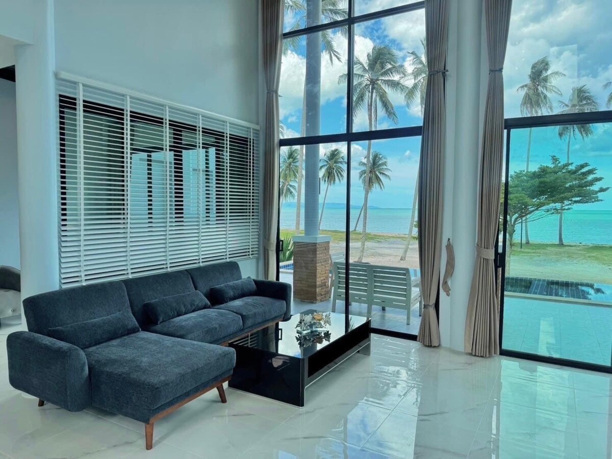 A modern living area features large floor-to-ceiling windows, providing a view of the nearby beach and palm trees. A dark-colored sofa is situated in front of a sleek glass coffee table, enhancing the open and airy feel of the space.