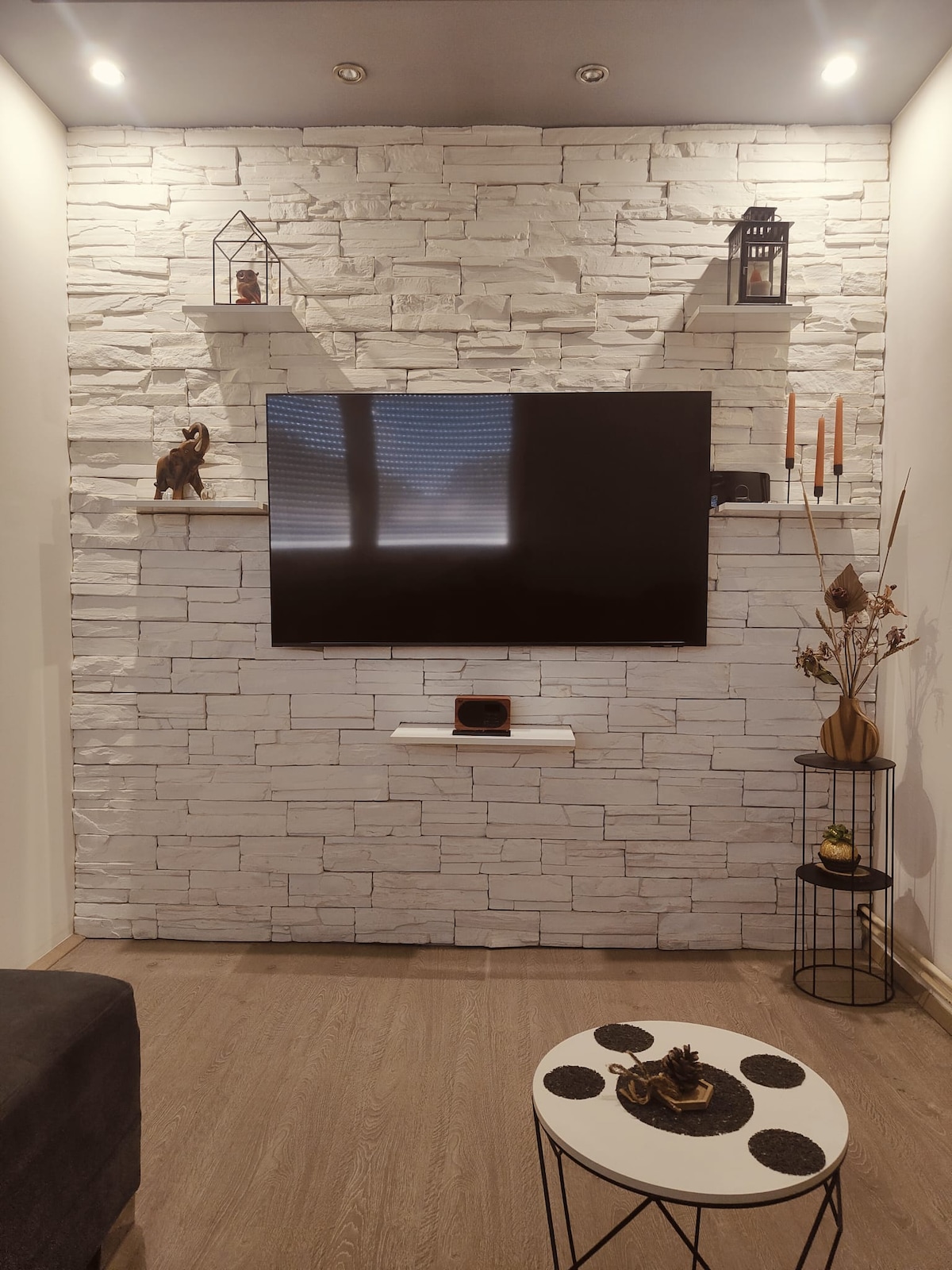 A modern living space features a white stone accent wall with decorative shelving. A flat-screen television is mounted onto the wall, and a small round coffee table, adorned with black coasters, sits in front of a dark sofa. Natural light filters gently through window blinds.