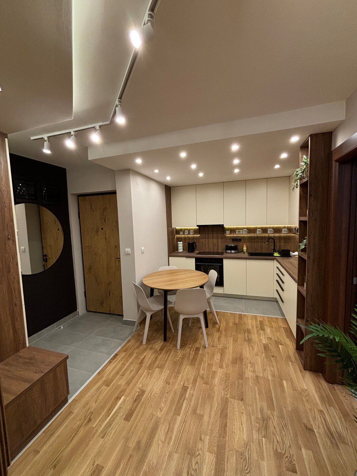 A modern kitchen area is presented with a round wooden dining table surrounded by four light-colored chairs. Soft overhead lighting brightens the space, which features a combination of wood and neutral cabinetry. The kitchen includes an array of appliances seamlessly integrated into the design.