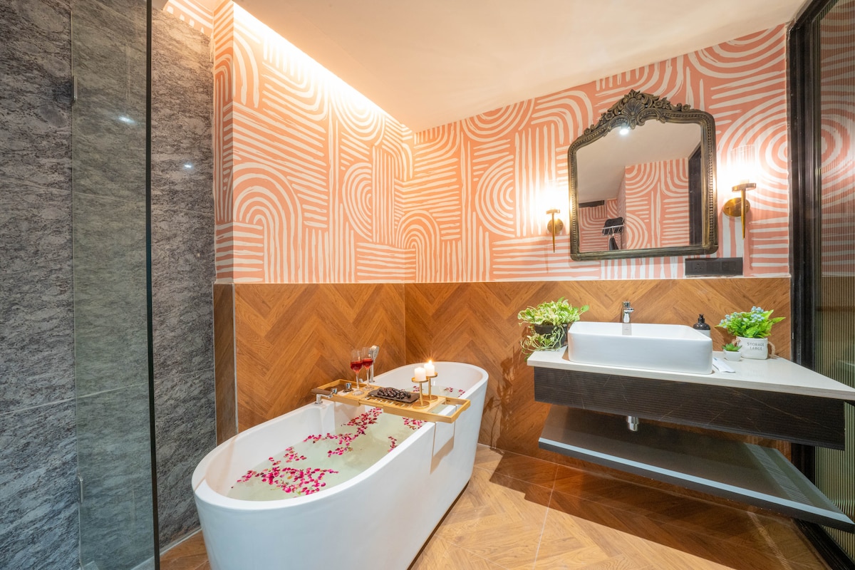 An elegant bathroom features a freestanding bathtub adorned with floating rose petals, complemented by a wooden tray with glasses. Modern fixtures are paired with a stylish mirror, and a decorative plant adds a touch of greenery. Textured walls create a contemporary ambiance.