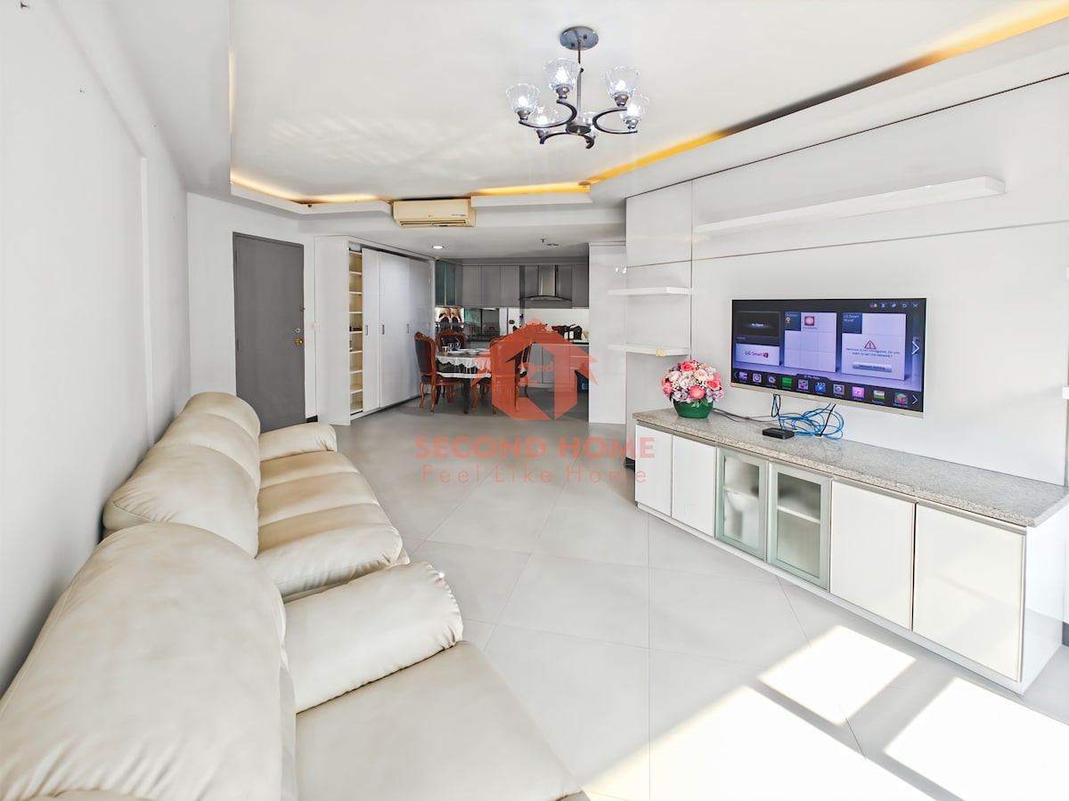 A bright living area features a comfortable sectional sofa and a modern entertainment unit with a 45'' SMART TV. Natural light fills the space, and a small decorative arrangement is seen atop the unit. The open layout connects to the kitchen, enhancing accessibility.