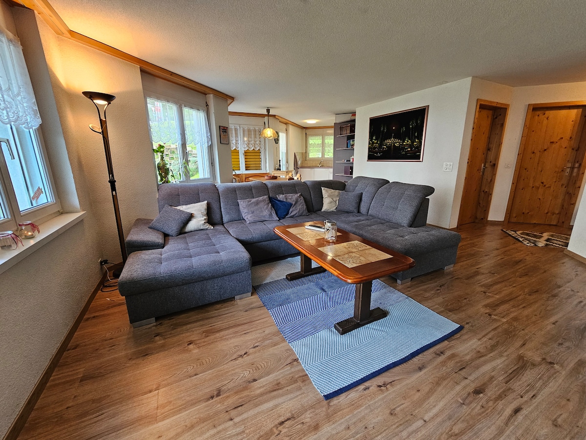 A spacious living area features a corner sofa in grey fabric, complemented by a wooden coffee table. Natural light fills the room through large windows adorned with sheer curtains. A woven rug is placed under the coffee table, adding texture to the wooden floor.