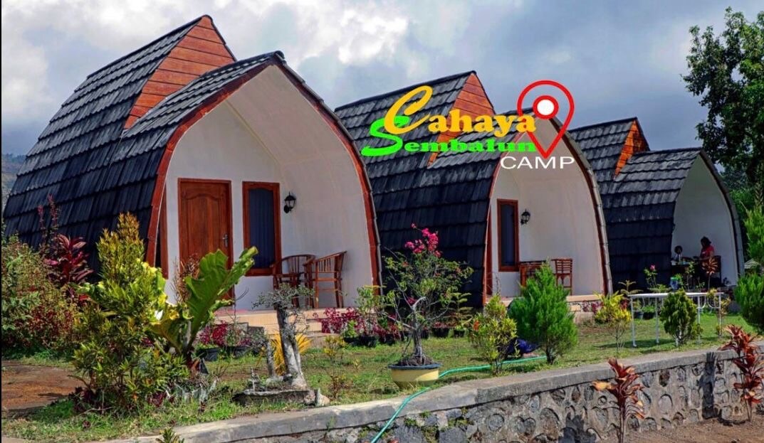 Several unique cottages are visible, featuring distinctive arched roofs and natural wooden doors. Lush greenery and colorful flowers surround the cottages, enhancing the tranquil outdoor space. A stone wall runs along the front, providing a defined area for the well-maintained landscape.