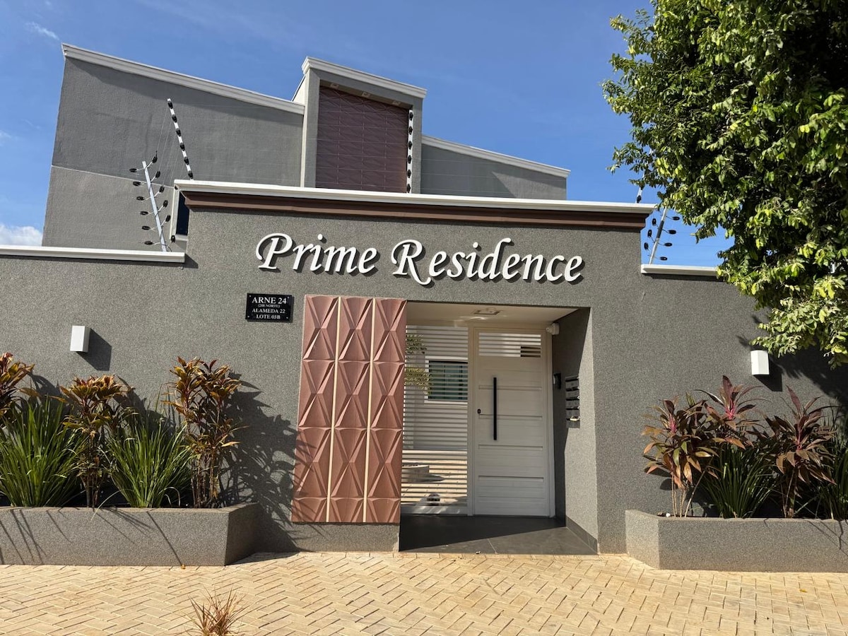 The entrance of Prime Residence is prominently displayed, featuring modern architecture with a mix of textures. The grey facade is complemented by lush greenery and decorative plants. A unique threshold design adds interest, while clear signage identifies the property.
