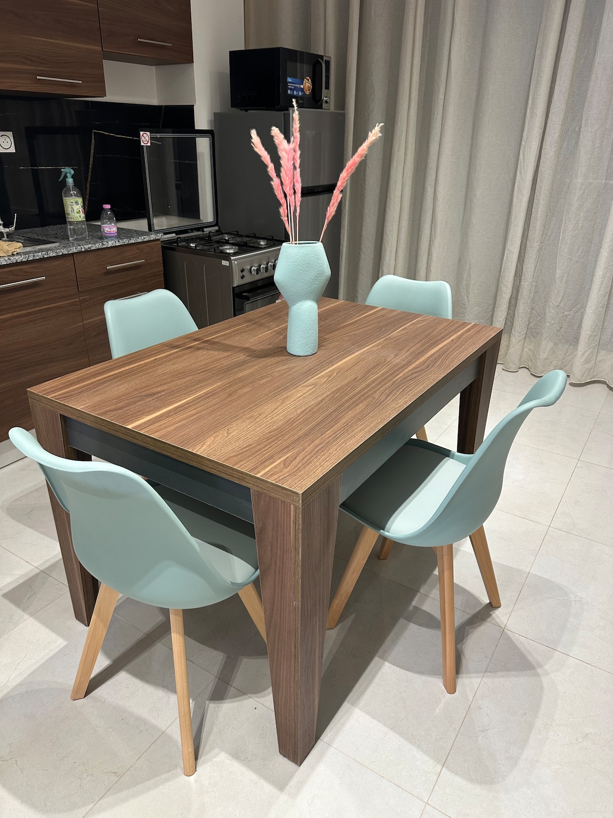 A modern dining area is displayed with a wooden table surrounded by four light blue chairs. A slender vase filled with decorative dried flowers is centered on the table. Soft, neutral drapes frame the space, complementing the sleek kitchen appliances in the background.