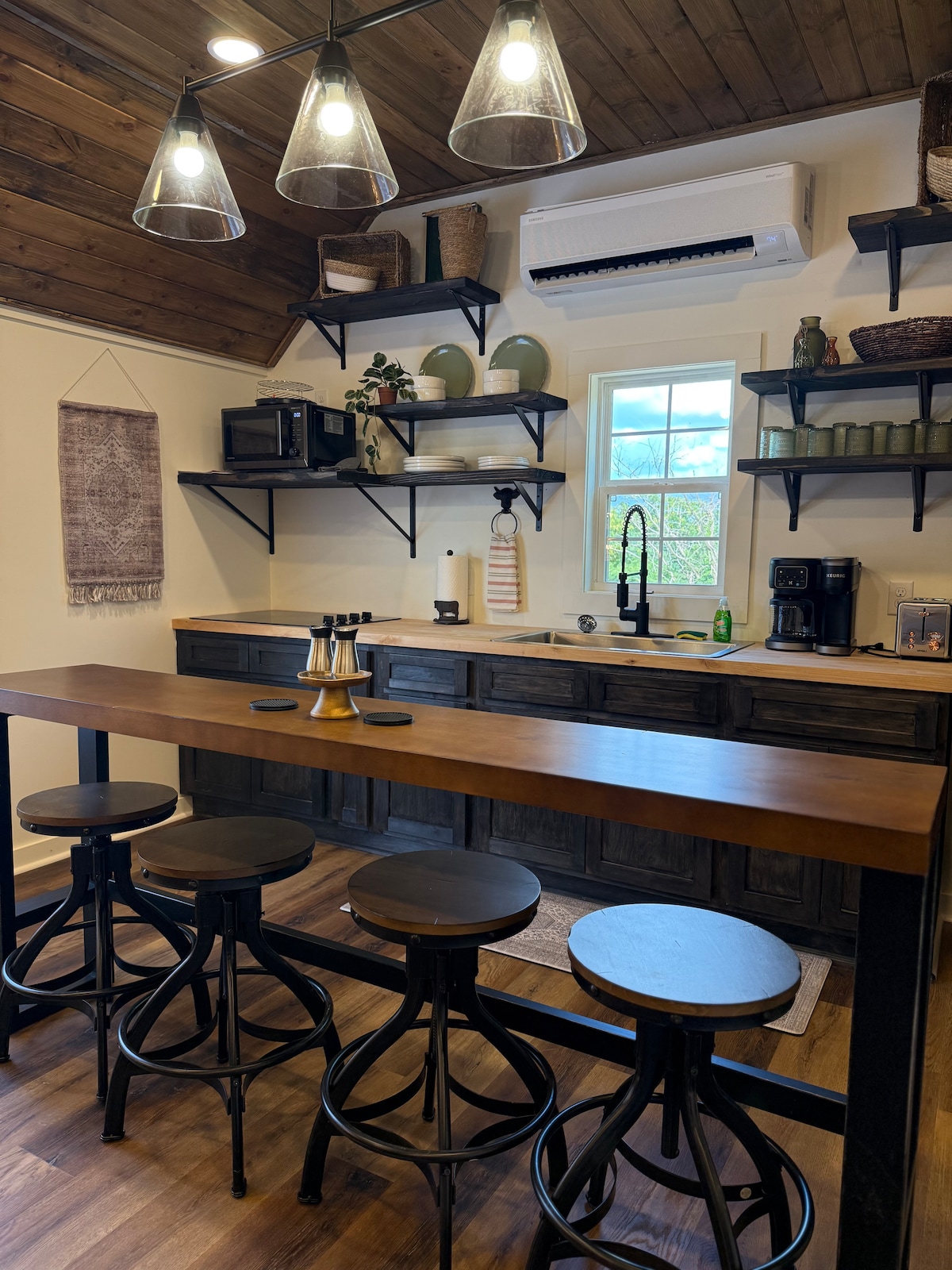 A modern kitchen area features wooden cabinetry with decorative open shelves displaying various kitchenware. A spacious wooden bar with four high stools is positioned near a window, providing natural light. Countertop appliances, a sink, and a microwave are visible, complementing the functional design.