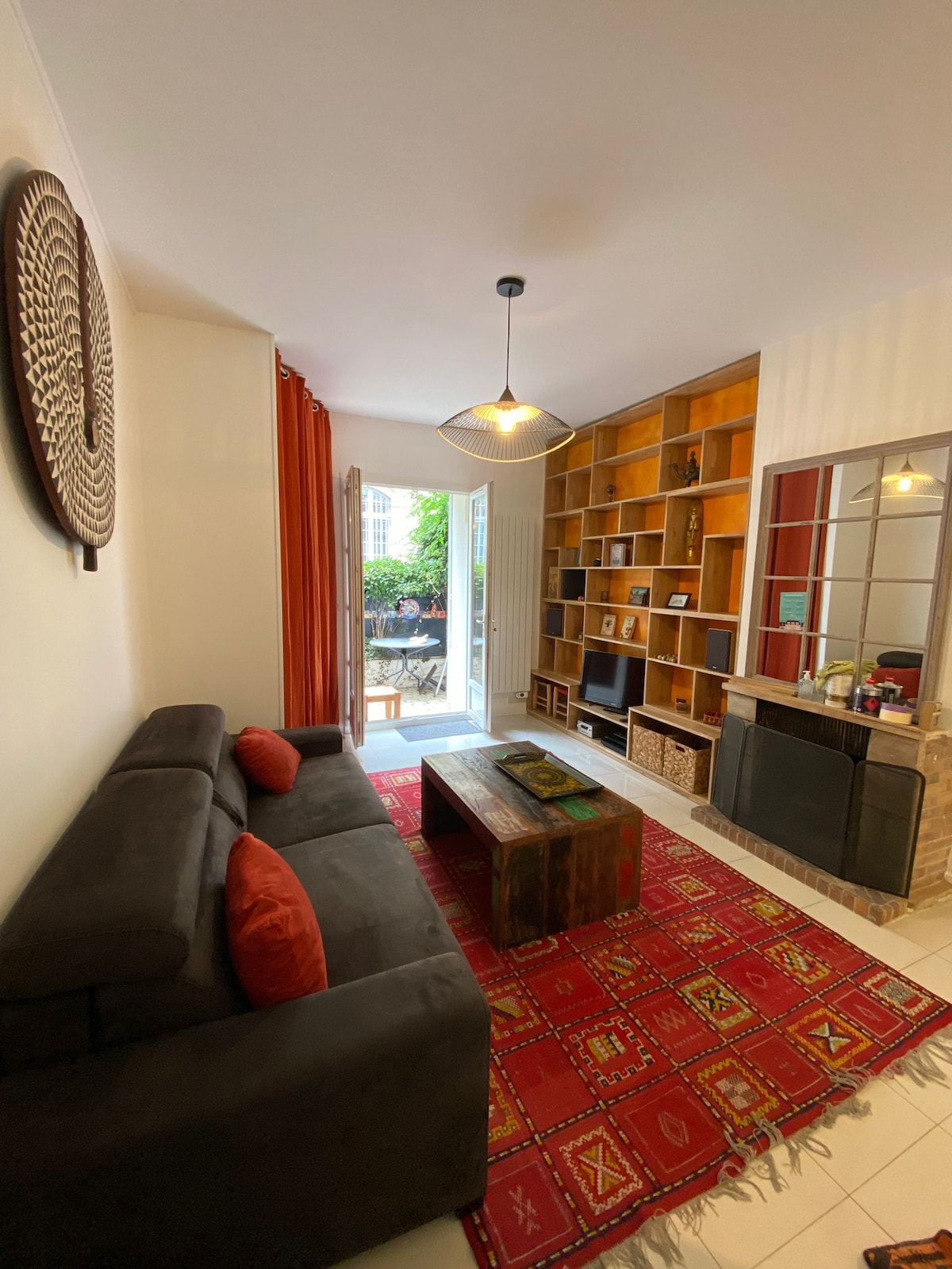 A comfortable living area features a dark sofa with decorative cushions and a wooden coffee table. A patterned rug covers the floor, and the space is enhanced by a decorative wall piece. Light enters through glass doors, leading to a private terrace filled with greenery.