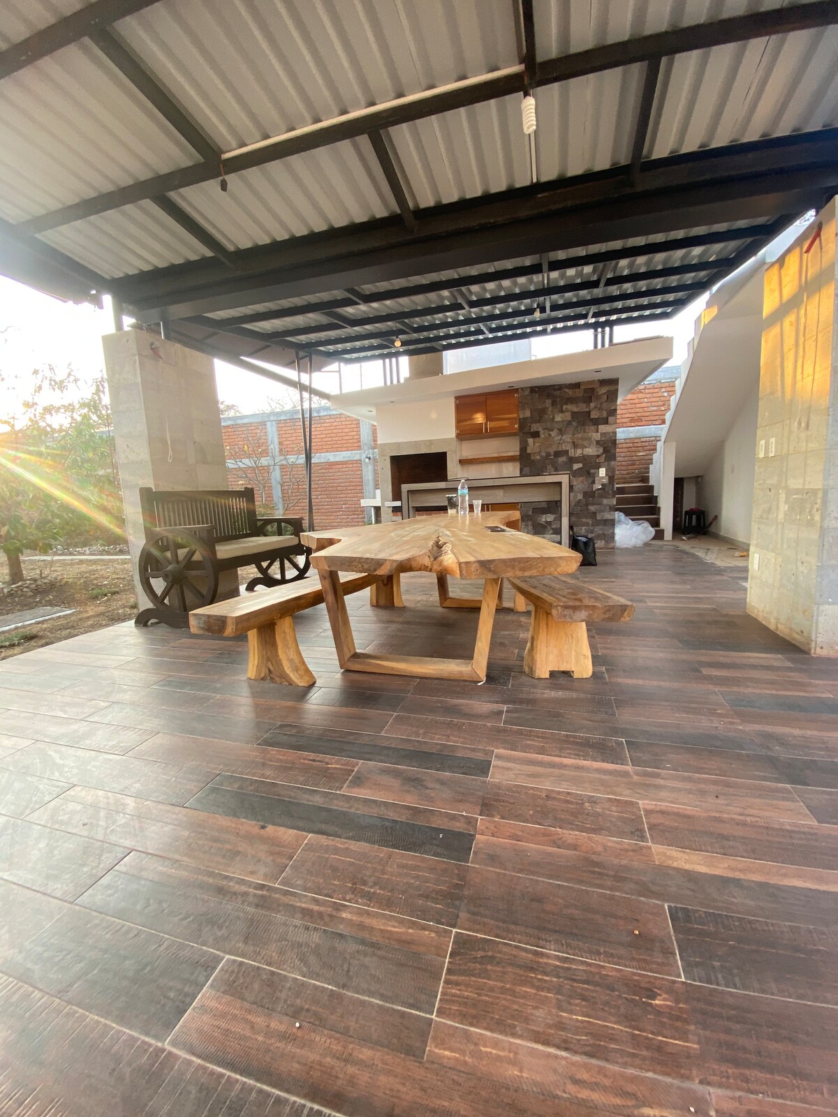 An outdoor dining area is showcased under a covered patio, featuring a large wooden table and matching benches. A stone fireplace is visible in the background, with a grill area nearby. Natural light filters in, enhancing the earthy tones of the space.