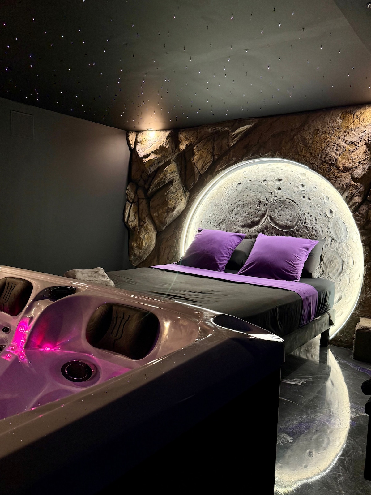 A cozy sleeping area is highlighted by a curved rock wall and a softly illuminated circular feature. The spacious bed is adorned with purple pillows and bedding. A sleek jacuzzi is positioned nearby, with subtle ambient lighting adding to the serene atmosphere.