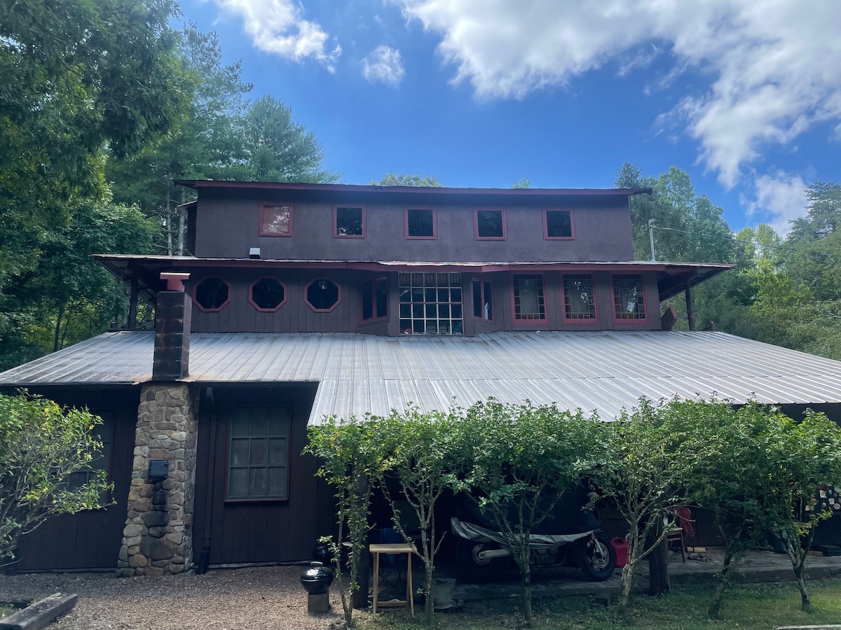 The lodge's exterior is characterized by a two-story structure with a unique design, featuring large round and rectangular windows. The roof has a metal surface, and the surrounding greenery includes shrubs and trees, creating a natural setting that complements the rustic architecture.