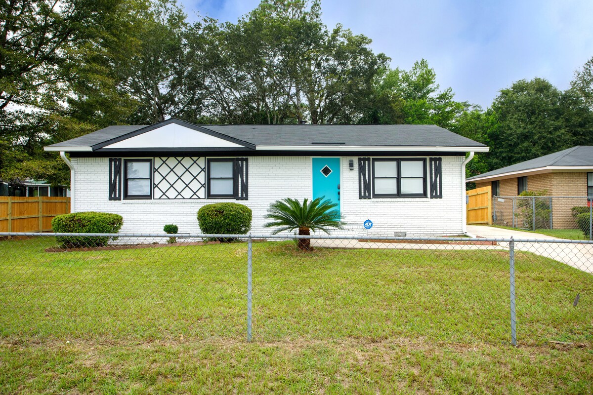 A single-story home is presented, featuring a white brick exterior complemented by bold black shutters and an eye-catching turquoise door. A manicured lawn is surrounded by a low fence, and a palm tree adds a touch of greenery to the front yard.