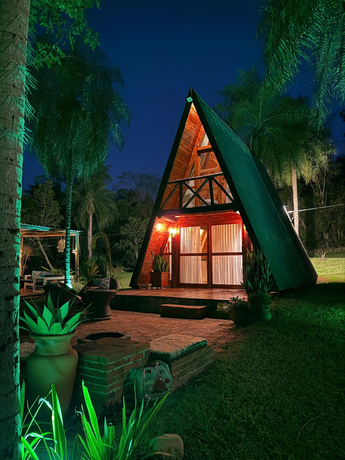 An A-frame chalet stands amidst lush greenery at night, softly illuminated by warm lights. The structure features large windows framed by wooden beams, while surrounding plants and trees create a serene environment. A pathway leads to the entry, enhancing the welcoming atmosphere.