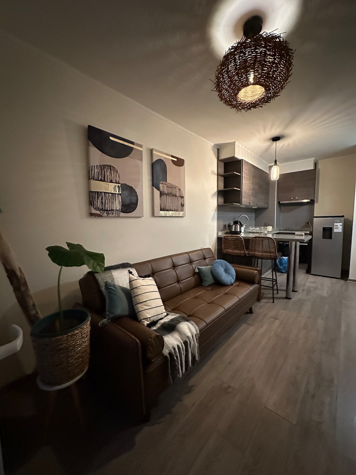 A modern living area features a sleek brown sofa adorned with decorative pillows. A woven light fixture hangs from the ceiling, casting soft illumination. Two abstract artworks adorn the wall, while a potted plant adds a touch of nature. In the background, the kitchen is visible with wood cabinetry.