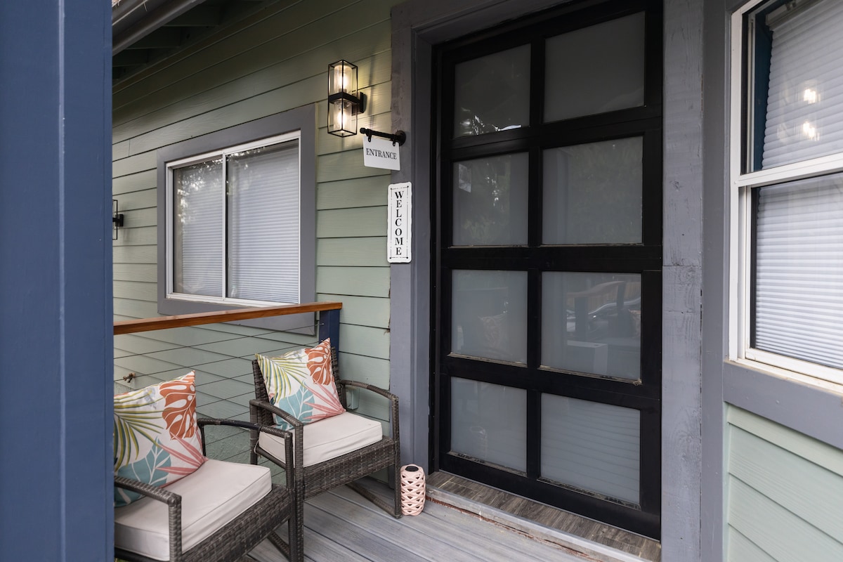 A welcoming entrance is visible, featuring a large black garage door and two comfortable chairs adorned with colorful cushions. A small decorative lantern adds charm, while a sign reads 'WELCOME.' Sunlight filters softly through the nearby windows.