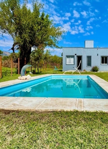 A refreshing swimming pool is surrounded by well-maintained grass and a few trees. Clear blue skies with scattered clouds are visible above, and a simple gray structure can be seen in the background, offering a serene outdoor space.